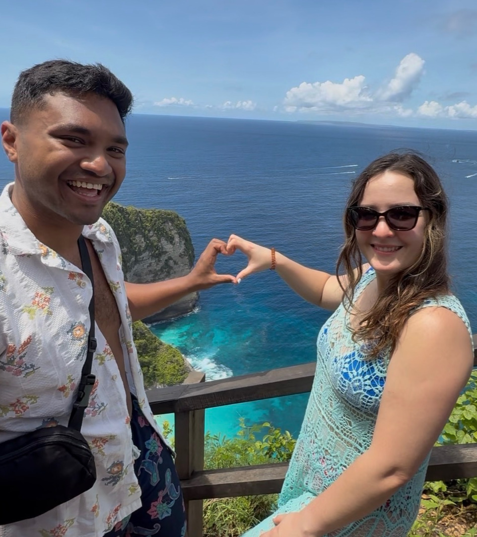 Giselle and Abhishek smiling and making a heart with their hands above the blue ocean